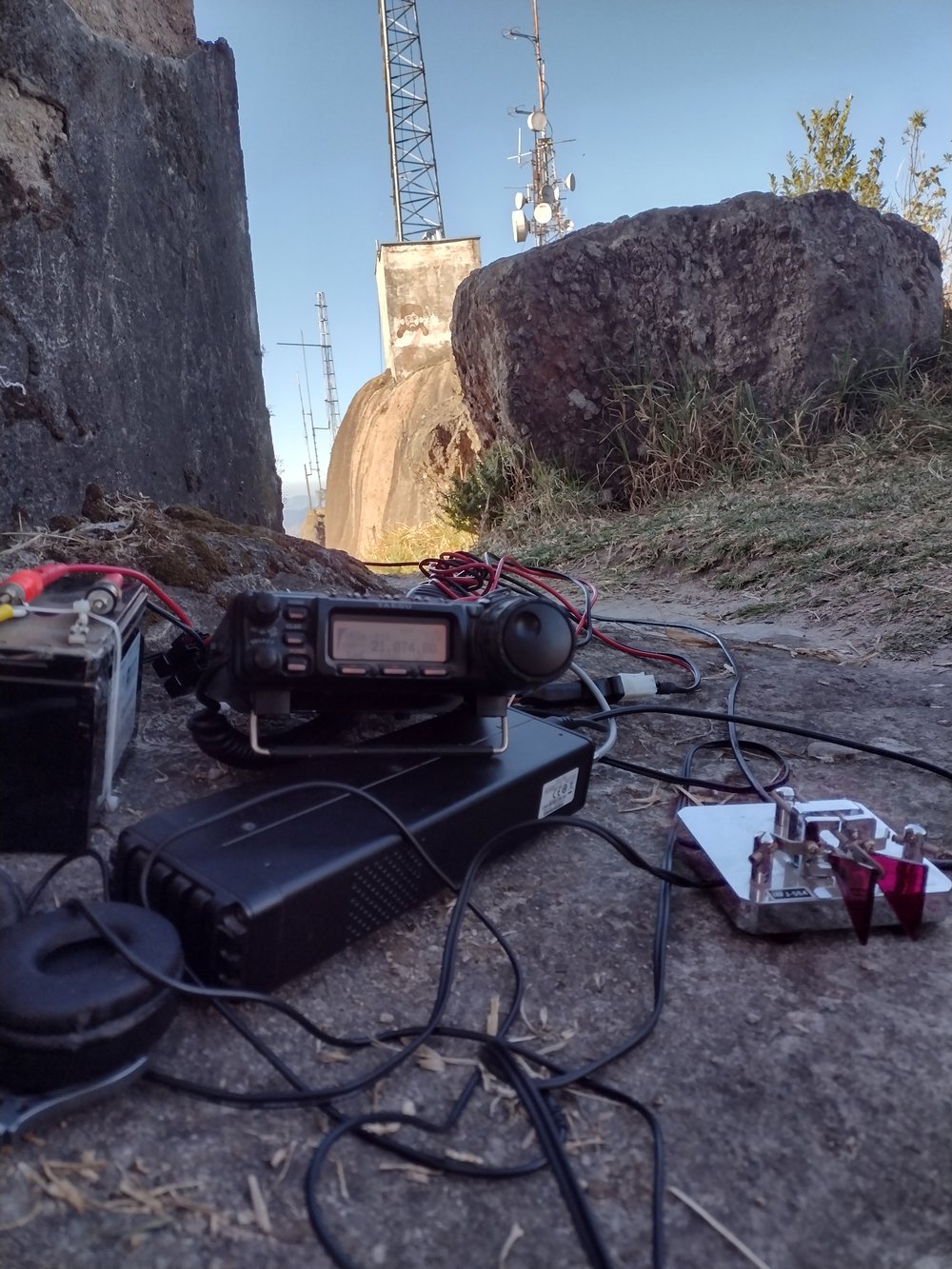 Figure 3 — A portable CW station on a mountaintop — a small HF transceiver, a battery, headphones, and a key on the rocks. You can work the world on a few watts and a wire. (Py4nf; Wikimedia Commons, CC BY 4.0.)