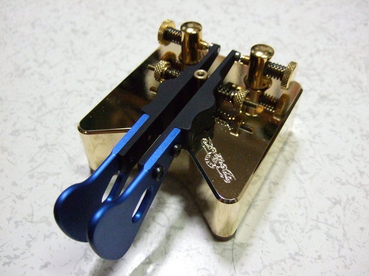 Figure 2 — A Begali Simplex — a jeweler-grade paddle with a mirror-polished solid-brass frame and base, gold thumbscrews, and blue-anodized levers. The input device as a keepsake. (しまでん / Shimaden, Wikimedia …