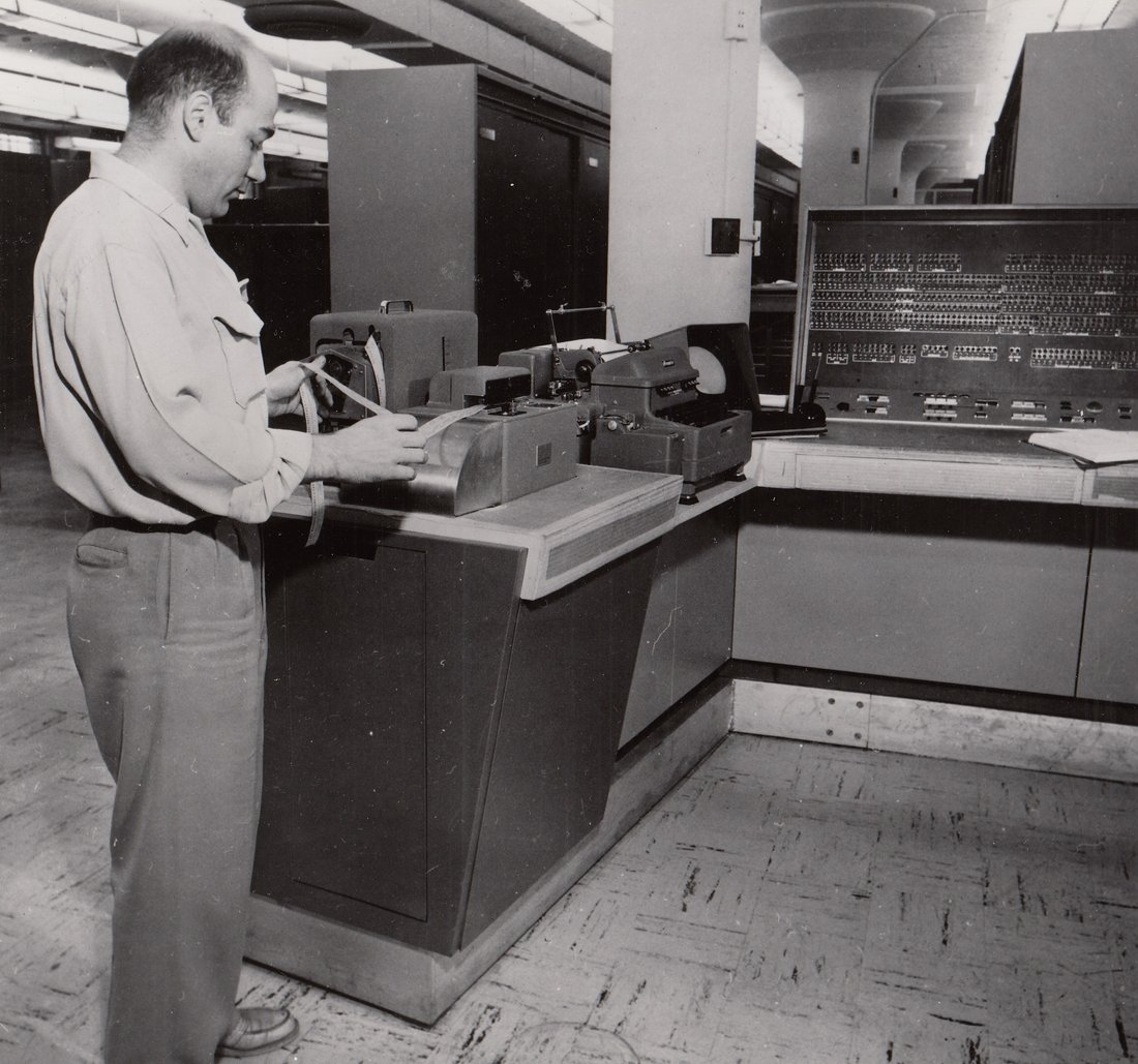 Figure 3 — A technician feeding a punched tape by hand into a reader at a computer console (a Univac 1105, 1950s) — the same paper ribbon that ran the torn-tape message networks also loaded the first computer…