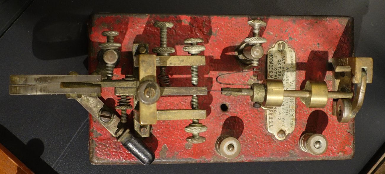 Figure 3 — Brass and bronze — the moving heart of a Vibroplex seen from above on its red crackle base. Even when the base is enameled, the mechanism gleams. (Daderot, National Electronics Museum; Wikimedia Co…