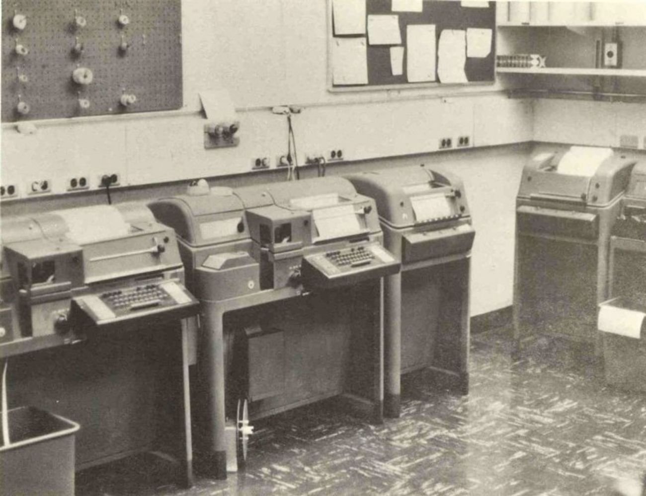 Figure 4 — A row of teletypes in a communications center (an ESSA forecast center, 1970) — the clattering rank of page printers that ran the news wires, the weather, the military traffic, and my father's shac…