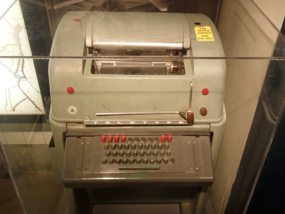 Figure 2 — A grey Teletype Model 28 with its red keys and domed cover — the ruggedized workhorse, with an exchangeable type box, that ran the military and financial wires. (Eric Fischer, New York Transit Muse…